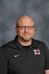 A man with glasses, a shaved head, and short beard wears a black collared shirt with a Nike logo and a red and white school emblem, posing in front of a gray background.