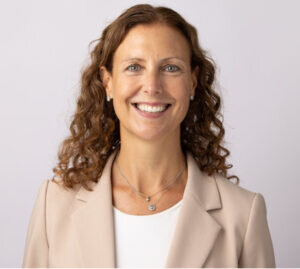 Smiling woman with curly brown hair wearing a beige blazer, white top, and layered necklaces, standing against a plain light background.
