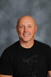 Man with black t-shirt smiles at the camera in front of a grey background