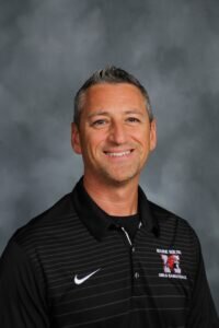 A man with short gray hair wearing a black Nike polo shirt with a Maine South Girls Basketball logo smiles in front of a gray background.