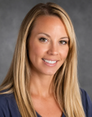 A woman with long blonde hair and a blue top smiles at the camera in front of a gray background.