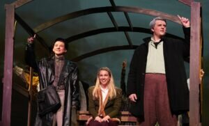 Three actors in period costumes stand on a stage set, with two standing and gesturing upward while one sits between them.