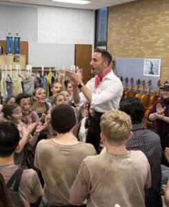 A male instructor in a white shirt and red neckerchief claps while facing a group of students in a classroom with music stands and trophies.