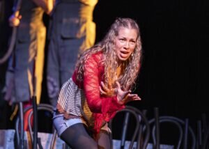 A performer with long, wavy hair sings expressively with her hands outstretched on a theater stage.