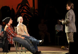 Two seated actors looking toward a third actor standing and holding a book on a stage.