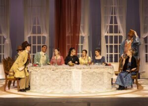 A group of actors in period costumes sit at a long dining table on a stage set.