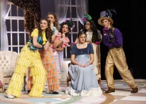 Six actors in eccentric, colorful period costumes perform on a stage, with one woman seated in a chair as the others look toward her.
