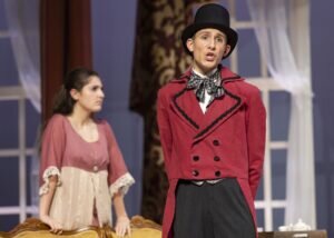 A male actor in a red tailcoat and top hat sings on stage while a woman in a period dress stands behind him.
