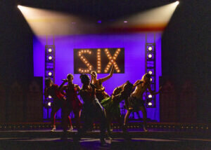 The cast of the musical SIX performing on stage in front of a large, illuminated sign reading SIX.