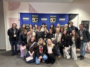 A group of students and adults stand in front of an Illinois Theatre Association 50th anniversary backdrop.
