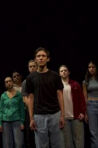 A group of six young people stands on a dark stage, looking forward with serious expressions.